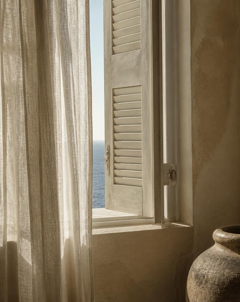 Linen curtain beside an open shuttered window with a sliver of sea — editorial photography
